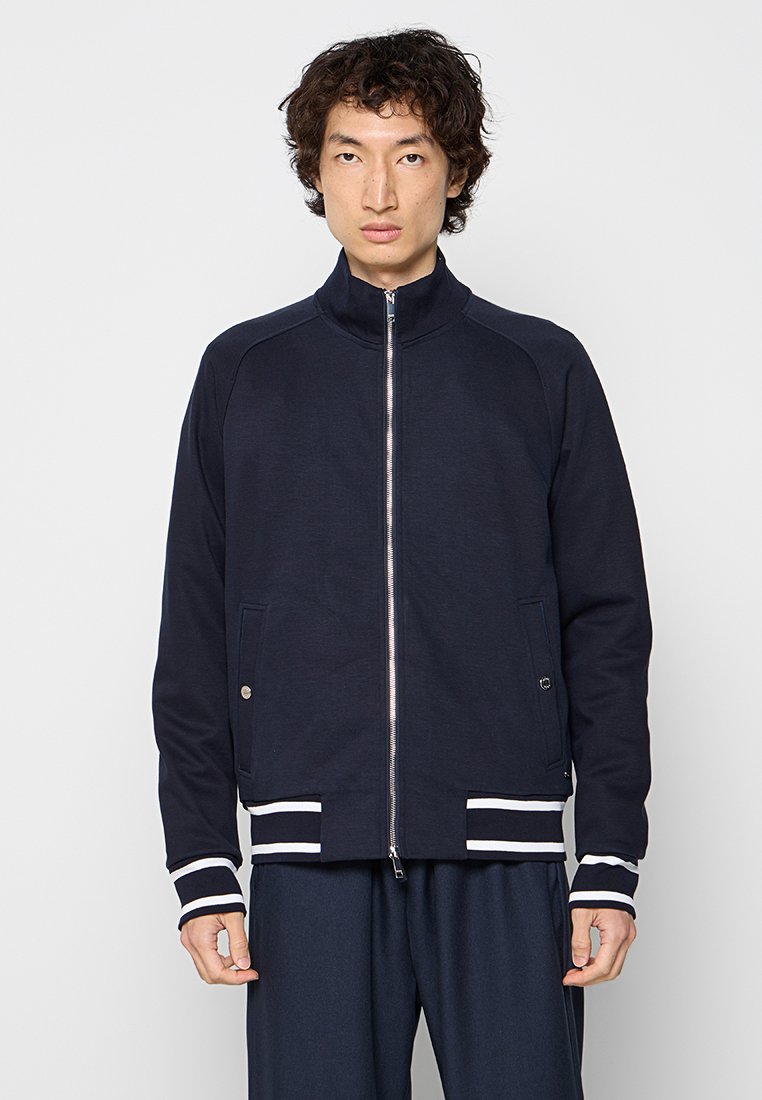 Granatowa kurtka zapinana na zamek z podwyższonym kołnierzem, ściągaczami na rękawach i u dołu, z białymi paskami, z dwoma bocznymi kieszeniami z zapięciami na zatrzask. Gładka tekstura.