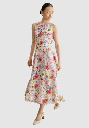 Femme portant une robe fleurie sans manches, longue jusqu'aux chevilles, avec des fleurs rouges, roses et violettes, associée à des chaussures beiges à talons blocs.