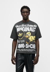 Νέος άνδρας που φοράει μαύρο υπερμεγέθες t-shirt Adidas με κινούμενο κίτρινο παπάκι και λευκό κείμενο, συνδυασμένο με μαύρη φόρμα Adidas με λευκές ρίγες.