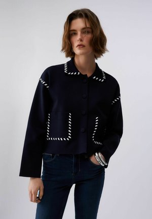 Žena oblečená v tmavomodrej zapínanej bunde s bielym šitím na golieri, vreckách a rukávoch, v kombinácii s tmavomodrými džínsami a striebornými náramkami.