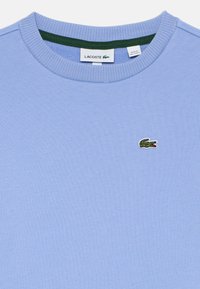 Felpa azzurra con colletto rotondo, etichetta del marchio Lacoste in verde e bianco e piccolo logo del coccodrillo verde sul petto.