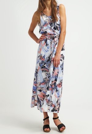Femme portant une robe sans manches longue jusqu'aux chevilles avec des motifs de feuilles colorées et des sandales noires à bride cheville, debout avec une main sur la hanche.
