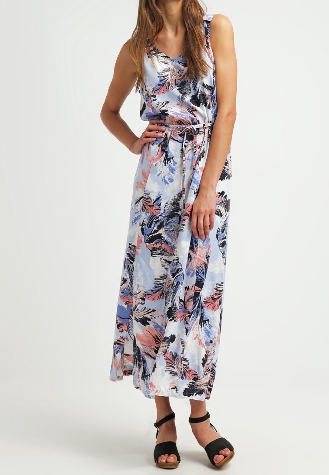 Femme portant une robe sans manches longue jusqu'aux chevilles avec des motifs de feuilles colorées et des sandales noires à bride cheville, debout avec une main sur la hanche.