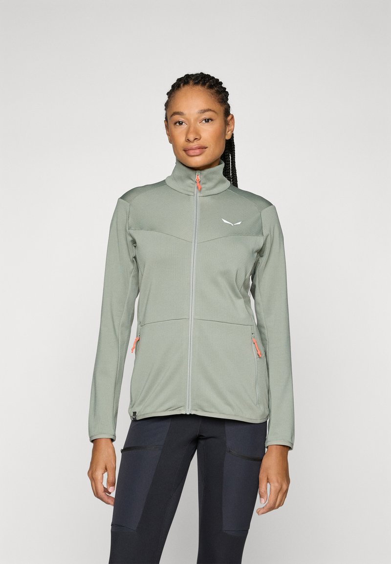 Lys grønn zip-up jakke med høy krage, teksturert stoff, oransje glidelåsdetaljer og sidelommer. Kombinert med svarte treningbukser.