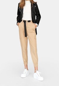 Beige cargobroek met elastische boorden, een riem met een zwarte gesp, gecombineerd met een witte coltrui en een zwart leren jack. Witte platform sneakers.