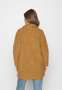 Brązowy płaszcz z futerka ekologicznego o miękkiej, teksturowanej powierzchni, z kołnierzem i luźnym krojem. Noszony na jasnoniebieskich dżinsach.