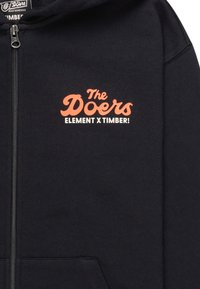 Zwarte rits hoodie van zacht materiaal, met op de linkerkant een oranje grafische tekst "The Doers ELEMENT X TIMBER!"