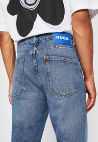 Jeans de mezclilla azul claro con costuras naranjas, mostrando una vista lateral de los bolsillos traseros y una etiqueta de marca azul con la inscripción "HUGO" en la cinturilla.