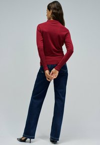 Mujer de pie con la espalda vuelta, vestida con una camiseta ajustada de manga larga roja, jeans rectos azul oscuro, tacones negros y pendientes de aro dorados.