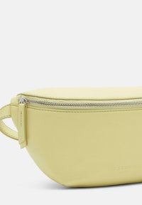 Żółta skórzana sakwa o zakrzywionym kształcie, z ozdobnym suwakiem w kolorze srebrnym i wytłoczonym logo. Materiał ma gładką teksturę i subtelny połysk.