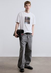 Λευκό μπλουζάκι με γραφικό σχέδιο, φαρδιά γκρι τζιν παντελόνια και μαύρα παπούτσια. Το μοντέλο κρατάει ένα μαύρο ρούχο τυλιγμένο. Απλό φόντο.