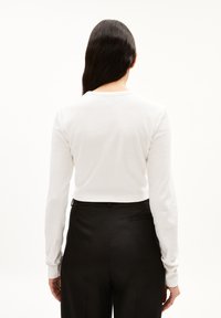 Langärmliges weißes Oberteil mit gerippter Textur und Rundhalsausschnitt, kombiniert mit schwarzen Hosen, die über eine Gesäßtasche verfügen.