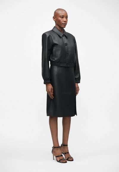 Crna kožna haljina s gornjim dijelom u stilu jakne s kragnom, elastičnim strukom i suknjom do koljena. Glatka tekstura, uski silueta, uparena s crnim štiklama.