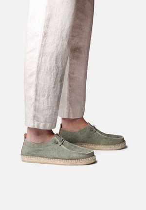 Person trägt hellbeige Hose und olivgrüne Espadrille-Casual-Schuhe aus Wildleder mit Seilsohlen und Schnürsenkeln.