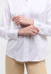 Weißes Baumwollhemd mit verdeckter Knopfleiste, langen Ärmeln und abgerundetem Saum. Der Stoff hat eine glatte Textur. Getragen mit beigen Hosen.