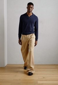 Navy long-sleeve polo shirt, beige wide-leg trousers, black footwear, standing on wooden floor, with a neutral-colored wall background.