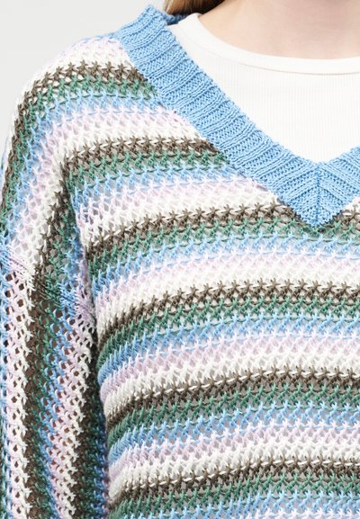 Gros plan sur un pull en maille ouverte multicolore avec des rayures horizontales bleu, vert, marron et blanc, porté sur un top blanc côtelé.