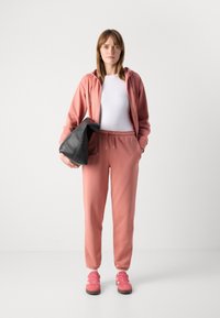 Ružová mikina a tepláky z mäkkého materiálu, doplnené o ružové tenisky. Jednotlivec drží čiernu kabelku. Minimalistický, ležérny dizajn.
