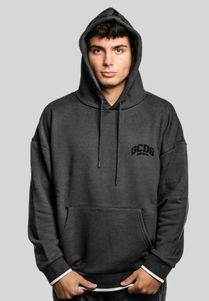 Giovane uomo che indossa una felpa con cappuccio GCDS grigio scuro, con il cappuccio alzato, in piedi davanti a uno sfondo grigio chiaro uniforme.