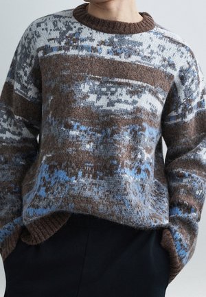 Personne portant un pull tricoté duveteux avec un motif pixellisé en marron, gris, blanc et bleu clair, mains partiellement dans les poches du pantalon noir.