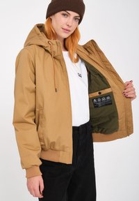 Tan gewatteerde jas met elastische manchetten en tailleband. Het interieur heeft een contrasterende groene voering en een verzorgingslabel. Het model draagt een wit shirt.