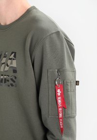 Felpa verde con testo camouflage sul petto e una tasca sulla manica con un portachiavi rosso "Rimuovere Prima del Volo".