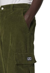 Mörkhyad person som bär olivgröna manchesterbyxor med en sidoficka och handen inskjuten i framfickan.