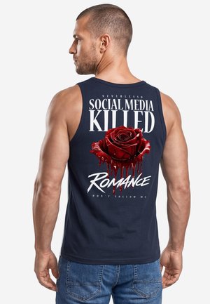 Muskelöser Mann mit kurzen Haaren, der ein schwarzes Tanktop trägt, auf dem eine tropfende rote Rose und der Text "Social Media Killed Romance" auf dem Rücken zu sehen sind.