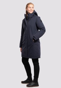 Parka azul marino hasta la rodilla con capucha, bolsillos laterales y cierre de cremallera. Fabricada con un material liso y resistente al agua. Botas de goma negras.