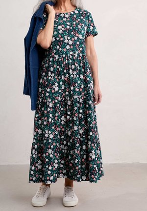 Robe midi fleurie en vert foncé avec des fleurs blanches et roses, manches courtes, design à volants, associée à des baskets blanches et une veste bleue.