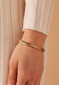 Gouden armband met een gladde, flexibele vormgeving en een golvend patroon, gedragen aan een hand tegen een lichtgekleurde gebreide stof als achtergrond.