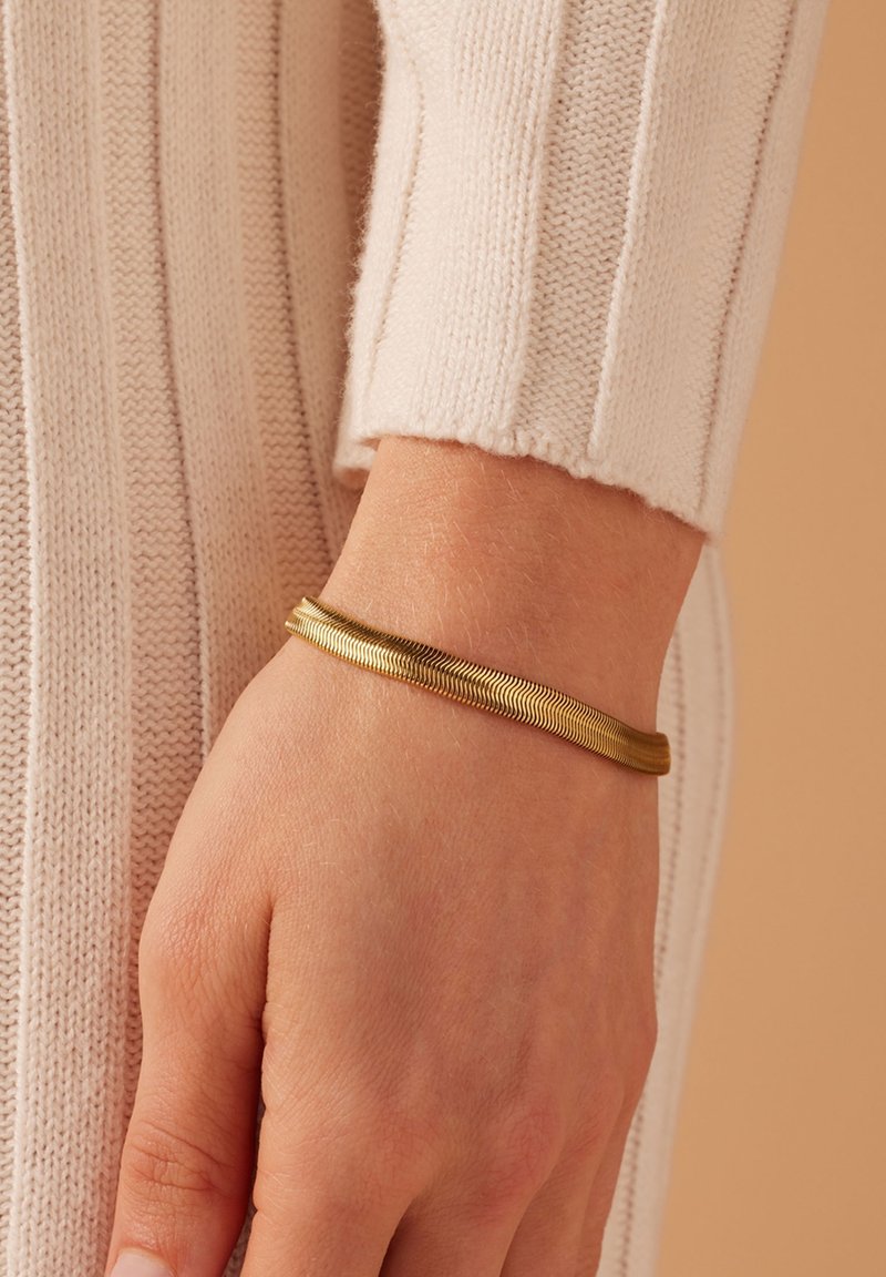 Gouden armband met een gladde, flexibele vormgeving en een golvend patroon, gedragen aan een hand tegen een lichtgekleurde gebreide stof als achtergrond.