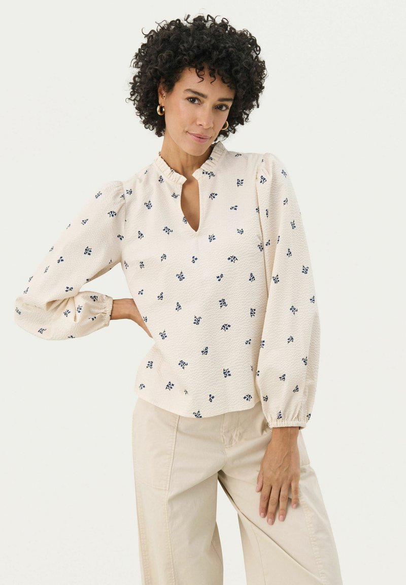 Blouse crème avec imprimé floral marine, décolleté en clef et manches bouffantes. Tissu texturé et poignets élastiques, associée à un pantalon beige.