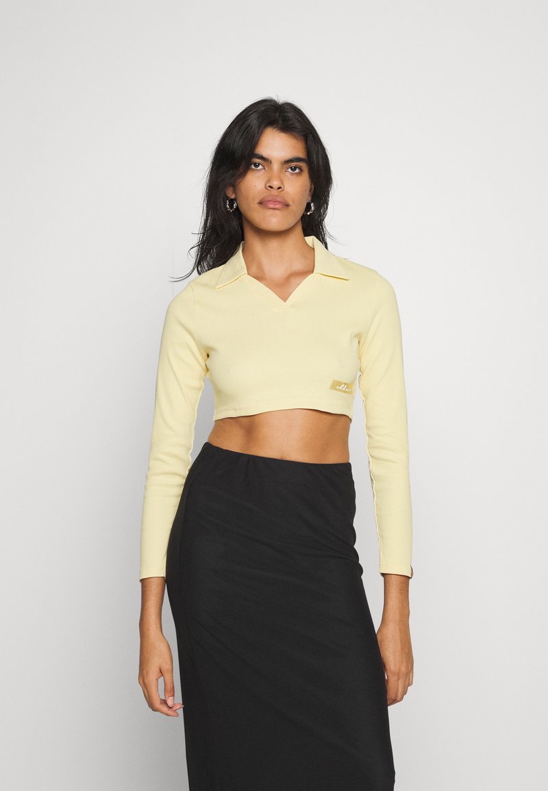 Top cropped amarelo de manga longa com um colarinho em V e logotipo bordado na bainha, combinado com uma saia preta. Textura suave, design justo.