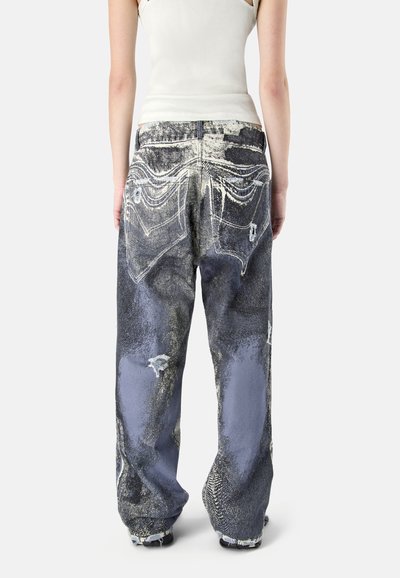 Personne portant un jean noir et gris délavé, ample et usé, ainsi qu'un débardeur blanc, vue de dos sur un fond uni.