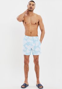 Blå blommiga badbyxor med dragsko, prydda med vita blommönster, tillsammans med svarta sandaler på en neutral bakgrund.