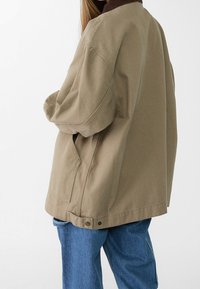 Übergroße braune Baumwolljacke mit hohem Kragen, seitlichen Taschen und glatter Textur, kombiniert mit blauen Denim-Jeans.