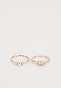 Deux alliances en or côte à côte. La bague de gauche présente une perle centrale flanquée de deux petites pierres; la bague de droite affiche un design ovale avec des pierres.