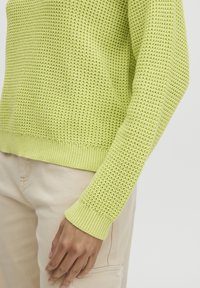 Strickpullover in hellem Limettengrün, mit gerippten Bündchen und Saum, strukturiertem Muster. Über hellbeigen Hosen getragen, die Detail der Ärmel zeigend.