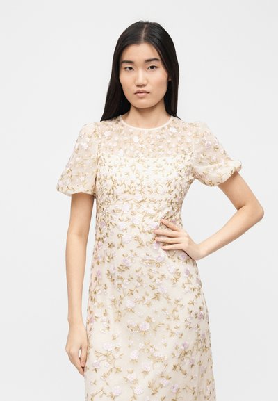 Νεαρή γυναίκα με μακριά σκούρα μαλλιά, ντυμένη με διαφανές, κεντητό μπεζ φόρεμα με λουλουδάτα σχέδια, στέκεται με το ένα χέρι στη μέση.