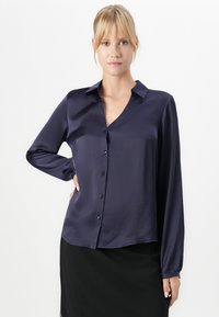 Blonde woman wearing a dark navy satin button-up blouse and black skirt, standing with one hand on her hip against a white background.