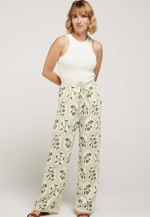 Femme portant un haut blanc sans manches et un pantalon large taille haute avec un motif floral et géométrique vert, debout les mains derrière le dos.