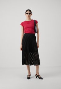 Red fitted t-shirt with logo, black pleated maxi skirt, sheer accents, black heeled slingback shoes, and round black sunglasses.