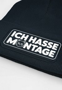 Black ribbed knit beanie featuring a white rectangular patch with the text "ICH HASSE MONTAGE" and a cat graphic. Smooth texture, casual design.