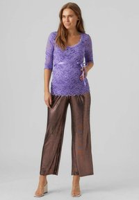 Fialový čipkovaný top s trojštvrťovými rukávmi, v kombinácii s lesklými hnedými širokými nohavicami. Obsahuje viazací detail v oblasti pása. Čisté pozadie.