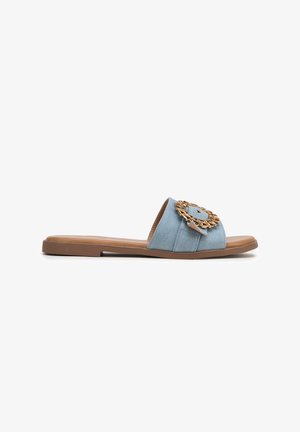Sandalo in denim azzurro chiaro con suola piatta, caratterizzato da un dettaglio circolare in tonalità dorata sulla fascia superiore. Texture liscia e design aperto.