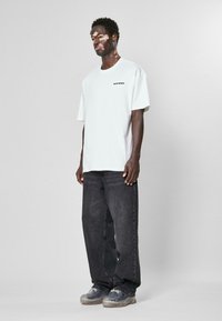 Tricou alb supradimensionat cu textul "SWIM SENSE." imprimat pe piept, asortat cu blugi negri largi și teniși translucizi gri.