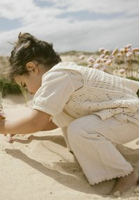 Niño vestido con un chaleco de ganchillo color crema sobre una camisa clara y pantalones beige con puntilla, agachado en la arena, interactuando con flores.