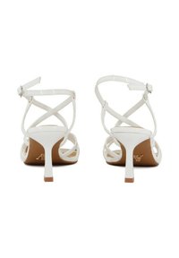 Sandali alti bianchi con cinturini incrociati e tacco sottile. Finitura lucida con suola color tan e un piccolo elemento metallico.