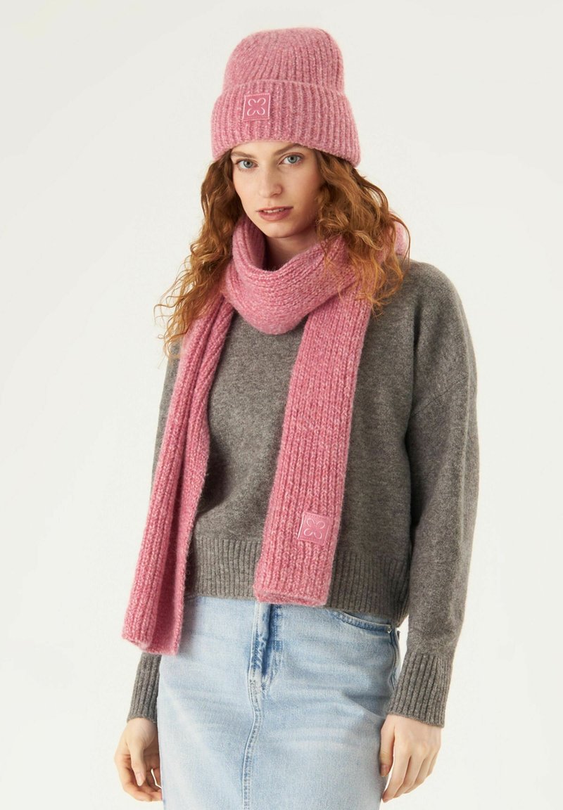 Pink knit beanie with ribbed texture and logo patch, matching pink scarf, gray sweater, and light blue jeans. Soft wool-like material.